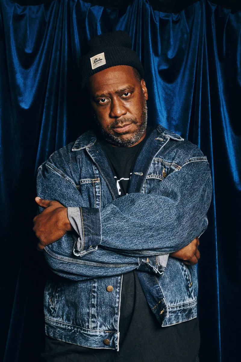 Robert Glasper in denim and a cap against a blue curtain at Blue Note London